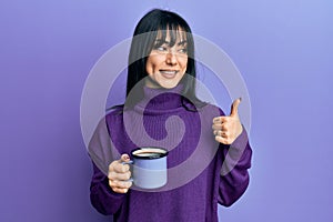 Young brunette woman with bangs drinking a cup of coffee smiling happy and positive, thumb up doing excellent and approval sign