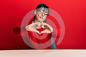 Young brunette woman with bangs wearing glasses sitting on the table smiling in love doing heart symbol shape with hands