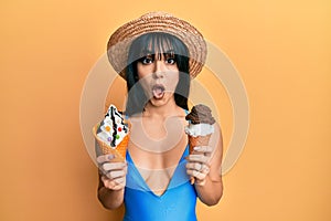 Young brunette woman with bangs wearing swimwear holding two ice cream cones afraid and shocked with surprise and amazed