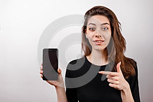 Young brunette woman in a black t-shirt smiling shows a phone on a white background. copyspase