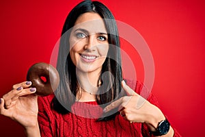 Young brunette woman with blue eyes eating chocolate doughnut over red isolated background with surprise face pointing finger to