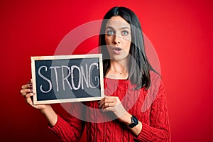 Young brunette woman with blue eyes holding blackboard with strong word message scared in shock with a surprise face, afraid and