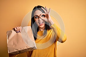 Young brunette woman with blue eyes holding delivery paper bag with food with happy face smiling doing ok sign with hand on eye