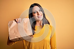 Young brunette woman with blue eyes holding delivery paper bag with food with a happy face standing and smiling with a confident