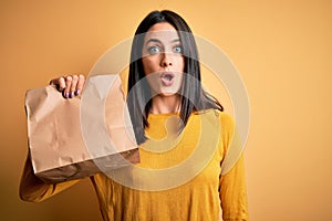 Young brunette woman with blue eyes holding delivery paper bag with food scared in shock with a surprise face, afraid and excited