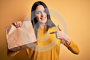 Young brunette woman with blue eyes holding delivery paper bag with food with surprise face pointing finger to himself