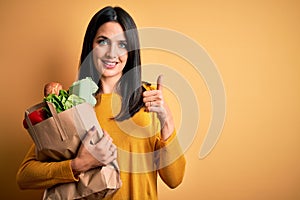 Young brunette woman with blue eyes holding healthy groceries in paper bag from supermarket happy with big smile doing ok sign,