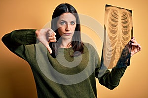 Young brunette woman with blue eyes holding xray of lungs over yellow background with angry face, negative sign showing dislike