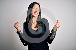 Young brunette woman with blue eyes wearing black casual sweater over white background very happy and excited doing winner gesture