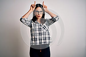 Young brunette woman with blue eyes wearing casual shirt and glasses over white background doing funny gesture with finger over