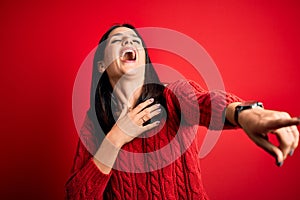 Young brunette woman with blue eyes wearing casual sweater over isolated red background laughing at you, pointing finger to the