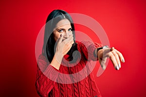 Young brunette woman with blue eyes wearing casual sweater over  red background laughing at you, pointing finger to the