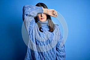 Young brunette woman with blue eyes wearing casual turtleneck sweater covering eyes with arm, looking serious and sad