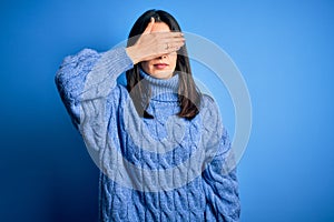 Young brunette woman with blue eyes wearing casual turtleneck sweater covering eyes with hand, looking serious and sad