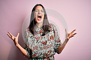 Young brunette woman with blue eyes wearing floral colorful dress over pink background crazy and mad shouting and yelling with