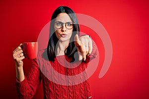 Young brunette woman with blue eyes wearing glasses and drinking a cup of coffee pointing with finger to the camera and to you,