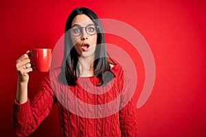 Young brunette woman with blue eyes wearing glasses and drinking a cup of coffee scared in shock with a surprise face, afraid and