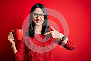 Young brunette woman with blue eyes wearing glasses and drinking a cup of coffee with surprise face pointing finger to himself