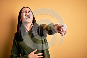 Young brunette woman with blue eyes wearing green casual sweater over yellow background laughing at you, pointing finger to the