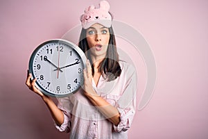 Young brunette woman with blue eyes wearing pajama holding big clock at night time scared in shock with a surprise face, afraid