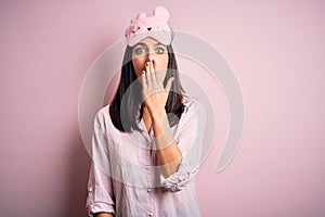 Young brunette woman with blue eyes wearing pink pajama and eye mask over pink background cover mouth with hand shocked with shame