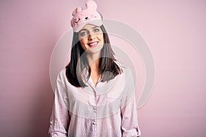 Young brunette woman with blue eyes wearing pink pajama and eye mask over pink background with a happy face standing and smiling