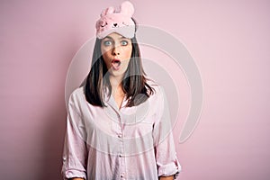 Young brunette woman with blue eyes wearing pink pajama and eye mask over pink background scared in shock with a surprise face,