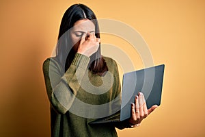 Young brunette woman with blue eyes working using computer laptop over yellow background tired rubbing nose and eyes feeling