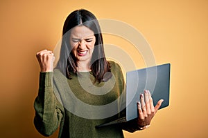 Young brunette woman with blue eyes working using computer laptop over yellow background very happy and excited doing winner