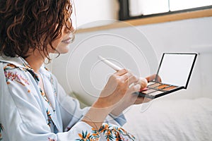 Young brunette woman in blue pajamas applies makeup with face sculpting palette sitting on bed at home
