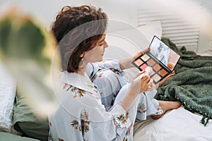 Young brunette woman in blue pajamas applies makeup with face sculpting palette sitting on bed at home