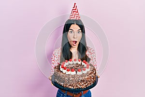 Young brunette woman celebrating birthday holding big chocolate cake afraid and shocked with surprise and amazed expression, fear