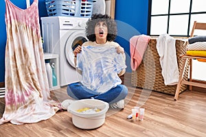 Young brunette woman with curly hair dyeing tye die t shirt and dress angry and mad screaming frustrated and furious, shouting
