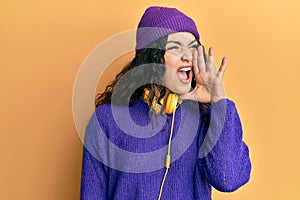 Young brunette woman with curly hair listening to music using headphones shouting and screaming loud to side with hand on mouth