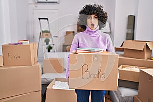 Young brunette woman with curly hair moving to a new home holding cardboard box in shock face, looking skeptical and sarcastic,