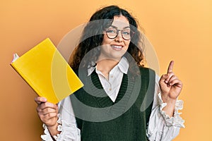 Young brunette woman with curly hair reading a book wearing glasses surprised with an idea or question pointing finger with happy