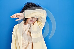 Young brunette woman with curly hair standing over blue background covering eyes with arm, looking serious and sad