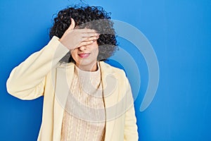 Young brunette woman with curly hair standing over blue background covering eyes with hand, looking serious and sad