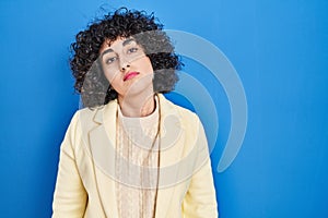Young brunette woman with curly hair standing over blue background looking sleepy and tired, exhausted for fatigue and hangover,