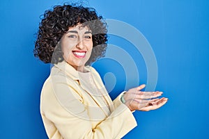 Young brunette woman with curly hair standing over blue background pointing aside with hands open palms showing copy space,
