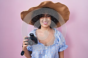 Young brunette woman with curly hair using reflex camera looking positive and happy standing and smiling with a confident smile