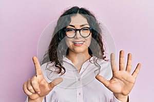 Young brunette woman with curly hair wearing casual clothes and glasses showing and pointing up with fingers number seven while