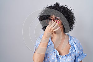 Young brunette woman with curly hair wearing glasses over isolated background smelling something stinky and disgusting,