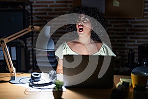Young brunette woman with curly hair working at the office at night angry and mad screaming frustrated and furious, shouting with