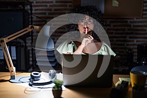 Young brunette woman with curly hair working at the office at night asking to be quiet with finger on lips
