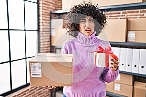 Young brunette woman with curly hair working at small business ecommerce holding present clueless and confused expression