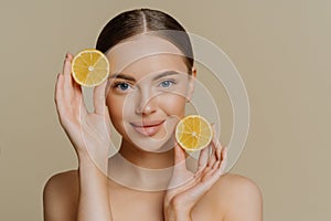 Young brunette woman with dark combed hair looks directly at camera holds two slices of lemons stands topless has