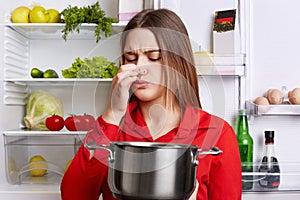 Young brunette woman with displeased expression smells spoiled soup in stew pan, feels musty smell at home kitchen, stands against