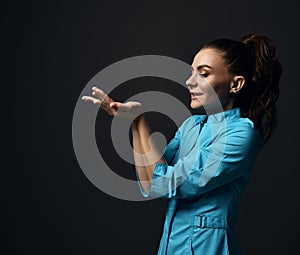 Young brunette woman doctor nurse in blue medical gown holds her open palms with copy space in front of her face