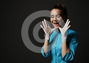 Young brunette woman doctor nurse in blue medical gown and latex gloves holds hands at face happy screaming